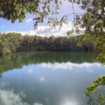 Tranquil cenote surrounded by lush greenery, reflecting the blue sky and clouds on its calm water surface.