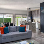 bright and spacious open-plan living area featuring a cozy gray sofa with colorful cushions, a dining space with large windows, and a modern kitchen in the background.