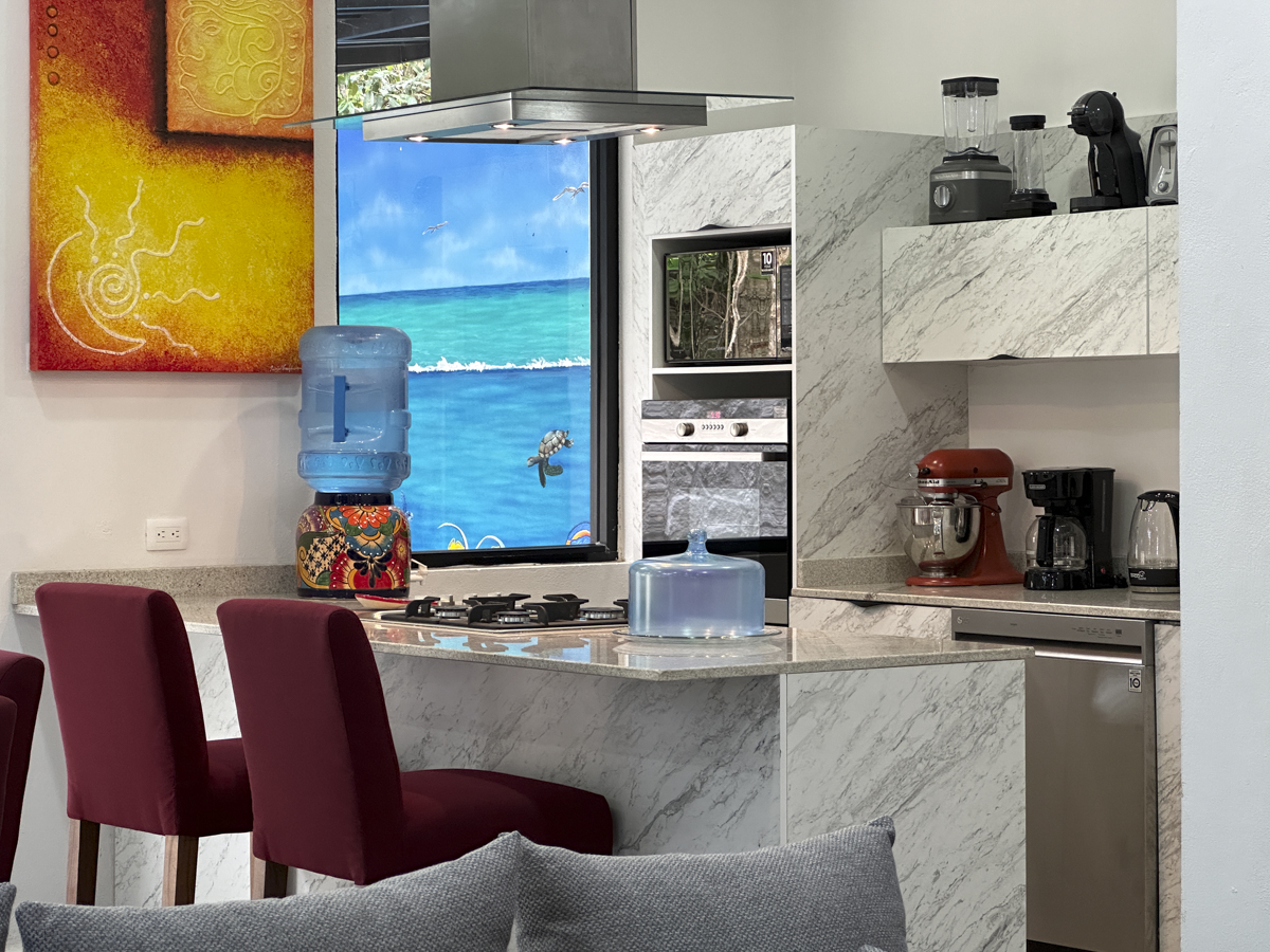 Modern kitchen with white marble countertops, stainless steel appliances, and red bar stools, complemented by vibrant artwork and a window mural depicting a beach scene.