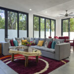 Spacious and modern living room with a large gray sectional sofa, colorful accent pillows, a natural wood coffee table, and floor-to-ceiling windows showcasing lush jungle views.