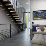 Modern loft-style interior featuring a sleek floating staircase with black metal railings, a cozy gray sectional sofa with colorful pillows, and framed artwork inspired by traditional Mexican culture.