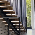 Close-up of a modern floating staircase with wooden steps, black metal framing, and vertical rope railings, set against large windows with jungle views.