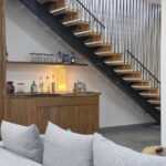 Stylish home bar area with a wooden counter and shelves stocked with glasses and spirits, set beneath a modern floating staircase with black metal railings.