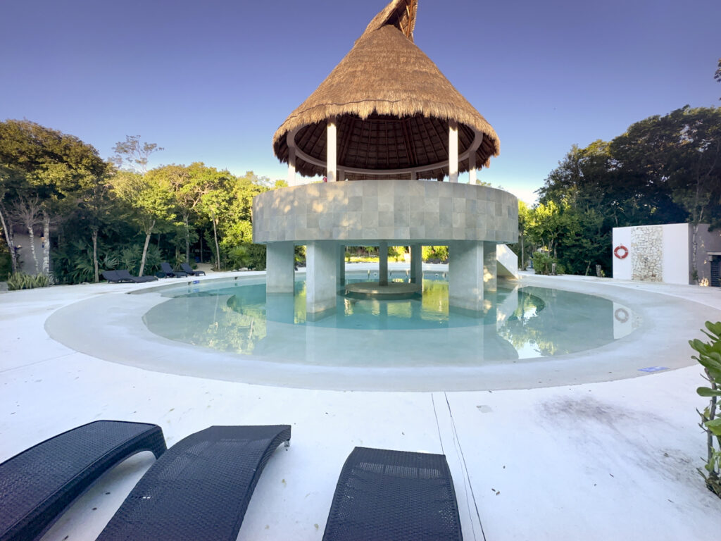 A luxurious pool with a thatched-roof palapa in a tropical setting, surrounded by lounge chairs and lush greenery.