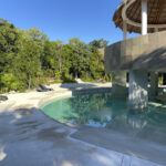 Luxurious swimming pool with a central palapa structure, surrounded by lounge chairs and lush jungle greenery under a bright blue sky.