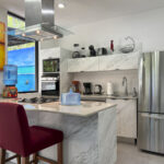 Modern kitchen with white marble countertops, stainless steel appliances, and red bar stools, complemented by vibrant artwork and a window mural depicting a beach scene.