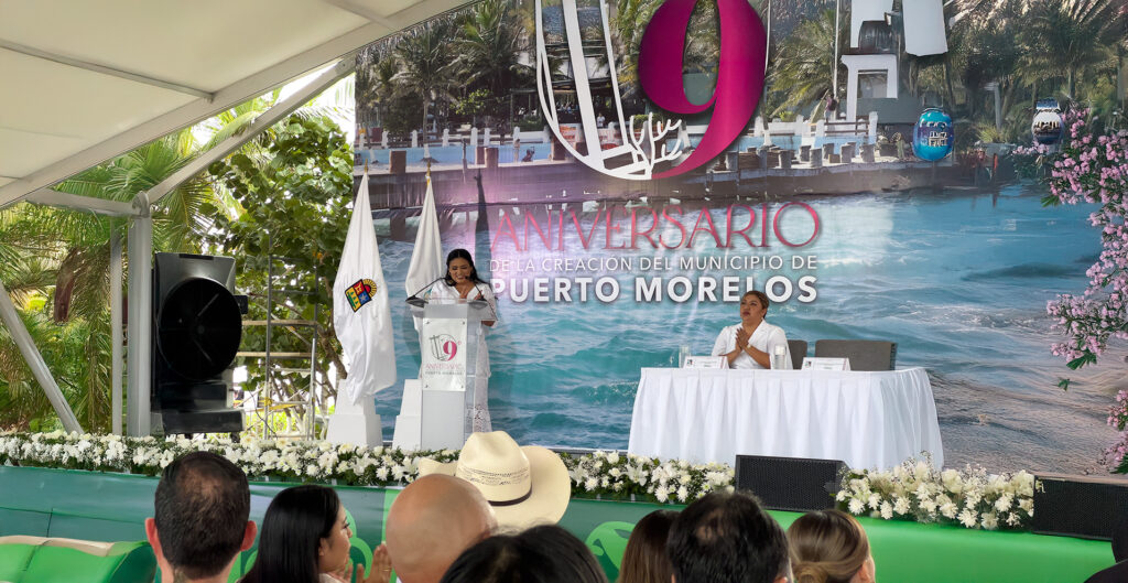 The mayor addresses the audience during the 9th-anniversary celebration of Puerto Morelos’ independence, with a festive backdrop and official decorations.