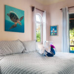 A bright and airy bedroom with a plush bed, ocean-themed artwork, and a private balcony overlooking tropical greenery.