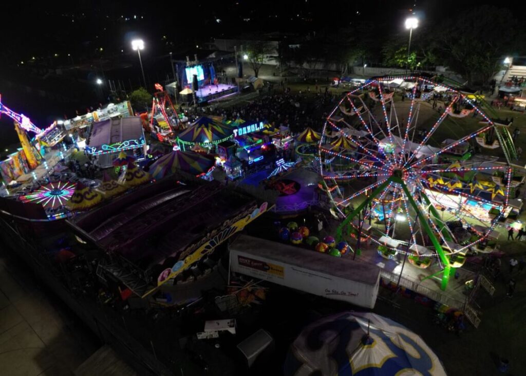 A vibrant aerial night view of the Puerto Morelos Carnaval fairgrounds, showcasing a Ferris wheel, colorful rides, game booths, and a stage lit up for live performances.