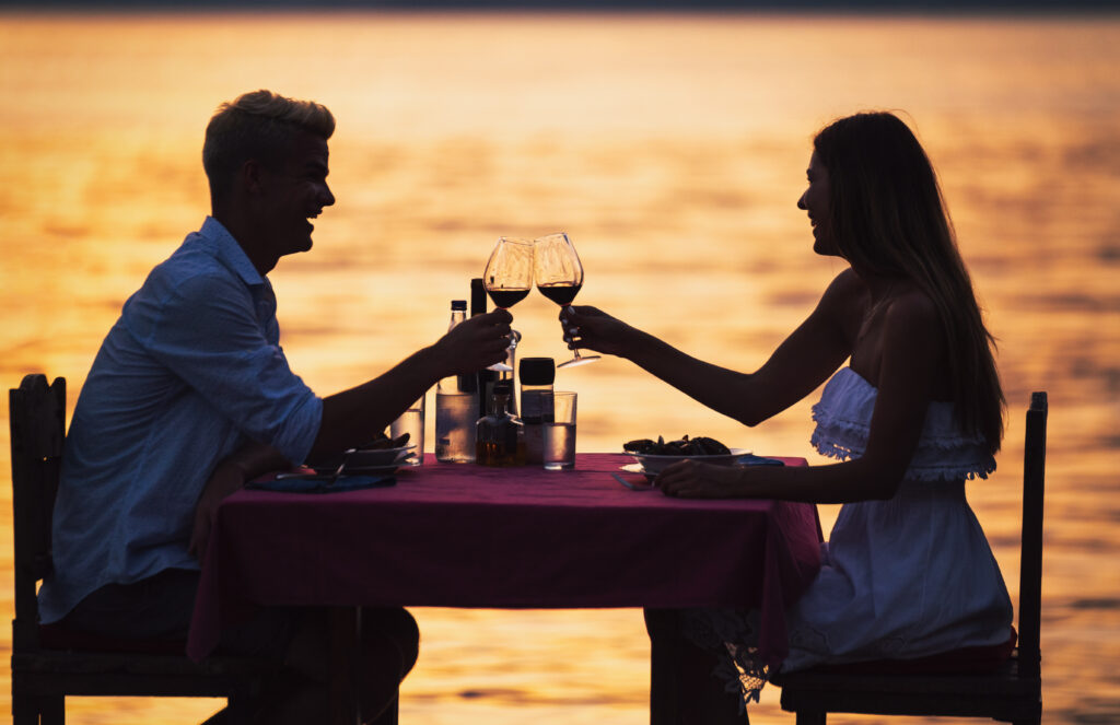 A couple enjoying a romantic dinner at sunset, clinking wine glasses by the water.