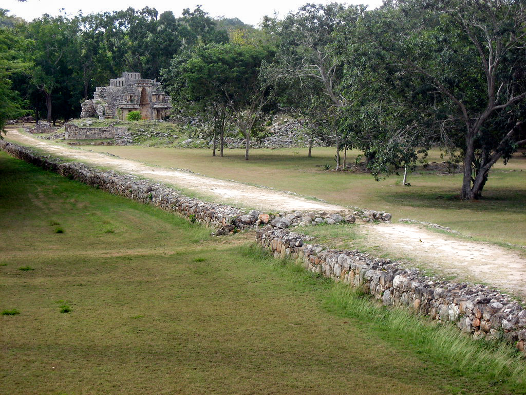 An elevated ancient Mayan white road, or sacbe, lined with stone walls, leading to a small Mayan ruin surrounded by trees and grassy fields in the Yucatán Peninsula.