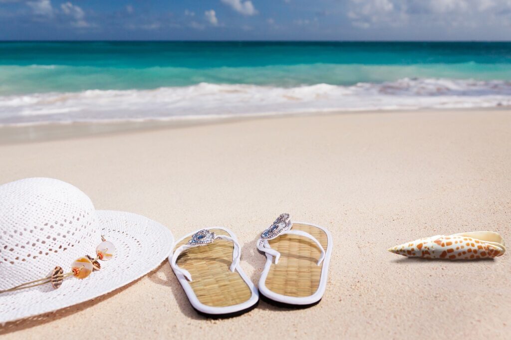 A white sun hat, stylish sandals, and a seashell resting on the sandy beach with turquoise ocean waves in the background.