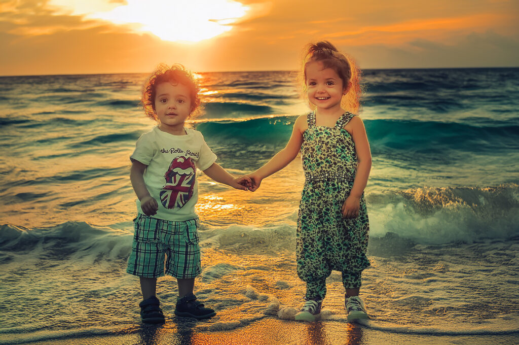 wo young children holding hands on the beach at sunrise, smiling as waves reach their feet.