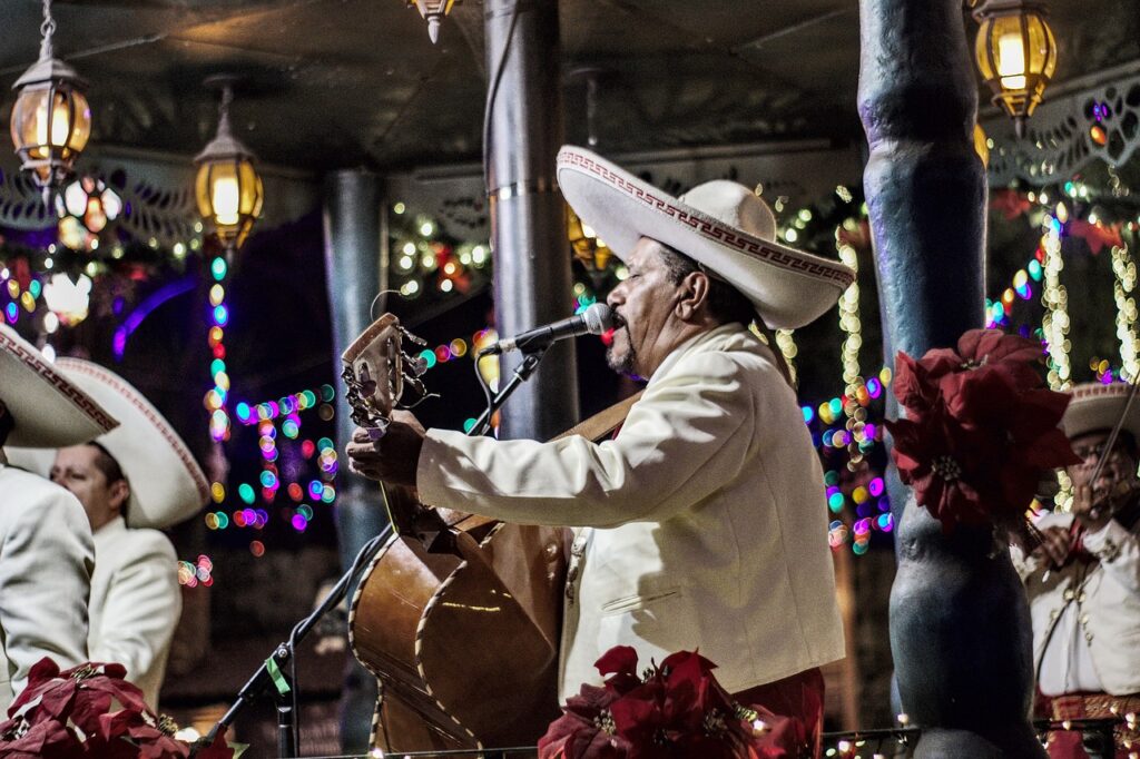 A mariachi musician in traditional white and red attire performs on stage with a guitar, surrounded by festive lights and decorations.