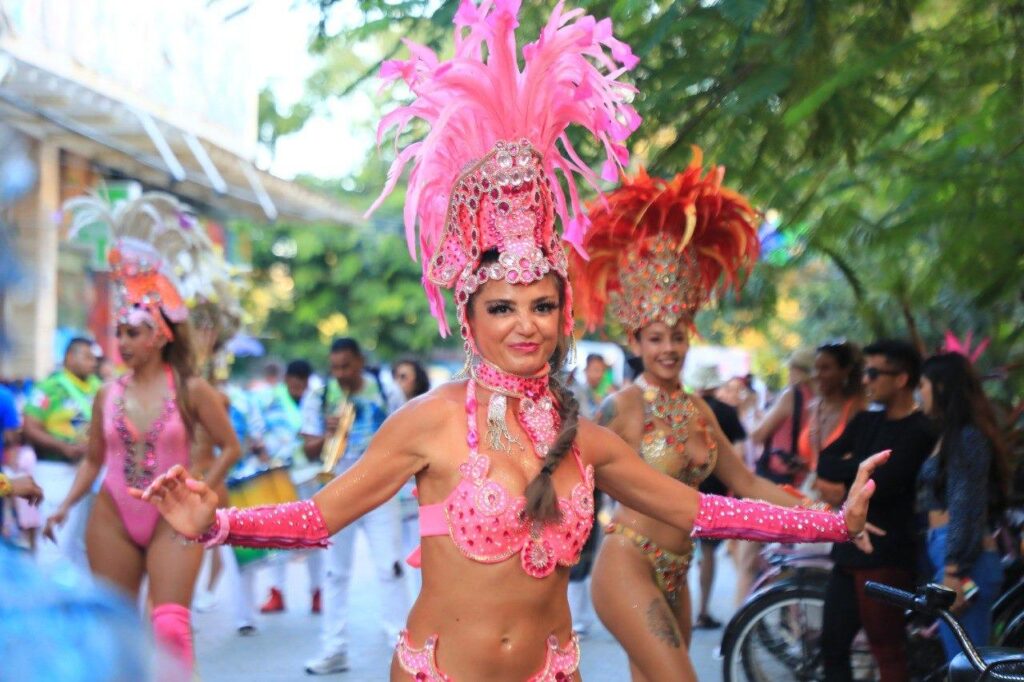 A vibrant Carnaval dancer in Puerto Morelos wearing a dazzling pink costume adorned with feathers, sequins, and intricate beading, smiling as she moves through the festive parade. Behind her, other performers in elaborate costumes and musicians add to the lively atmosphere.