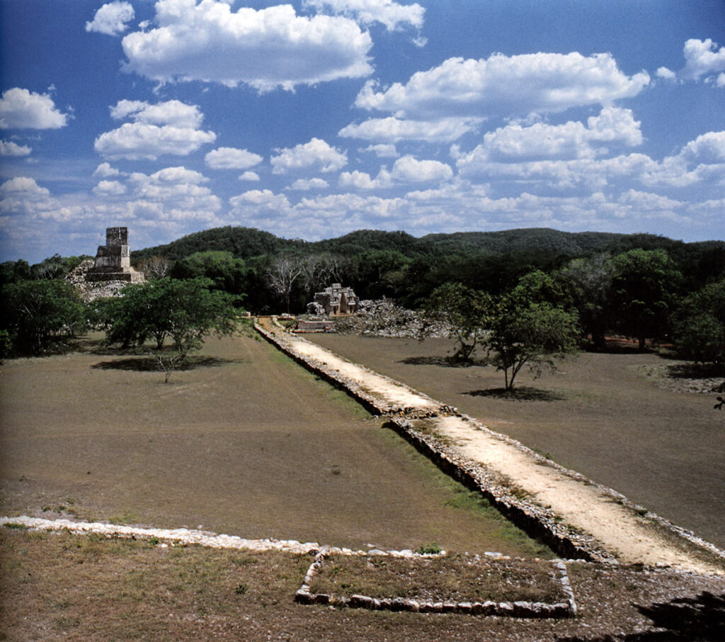 A wide, elevated ancient Mayan sacbe (white road) stretching through an open grassy area, leading to distant Mayan ruins surrounded by trees under a bright blue sky with scattered clouds.