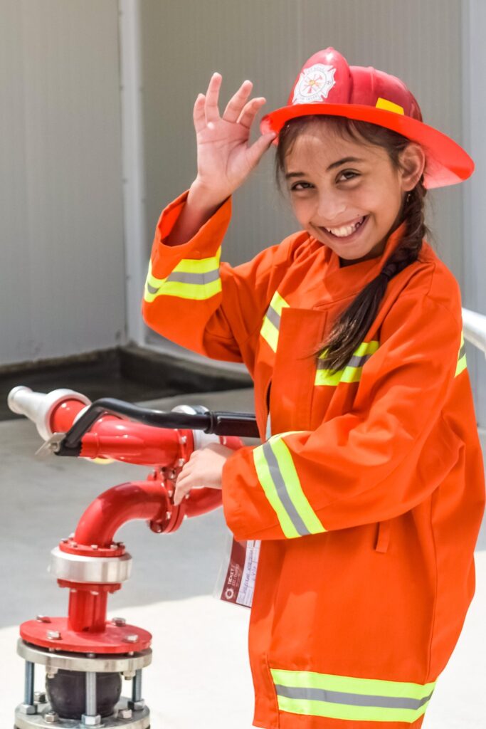 A smiling young girl in a bright orange firefighter uniform and red helmet poses with a fire hose valve, tipping her hat playfully.