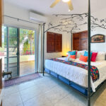 Casa Zarah bedroom with king bed, colorful pillows, canopy frame, and balcony view of palm trees.