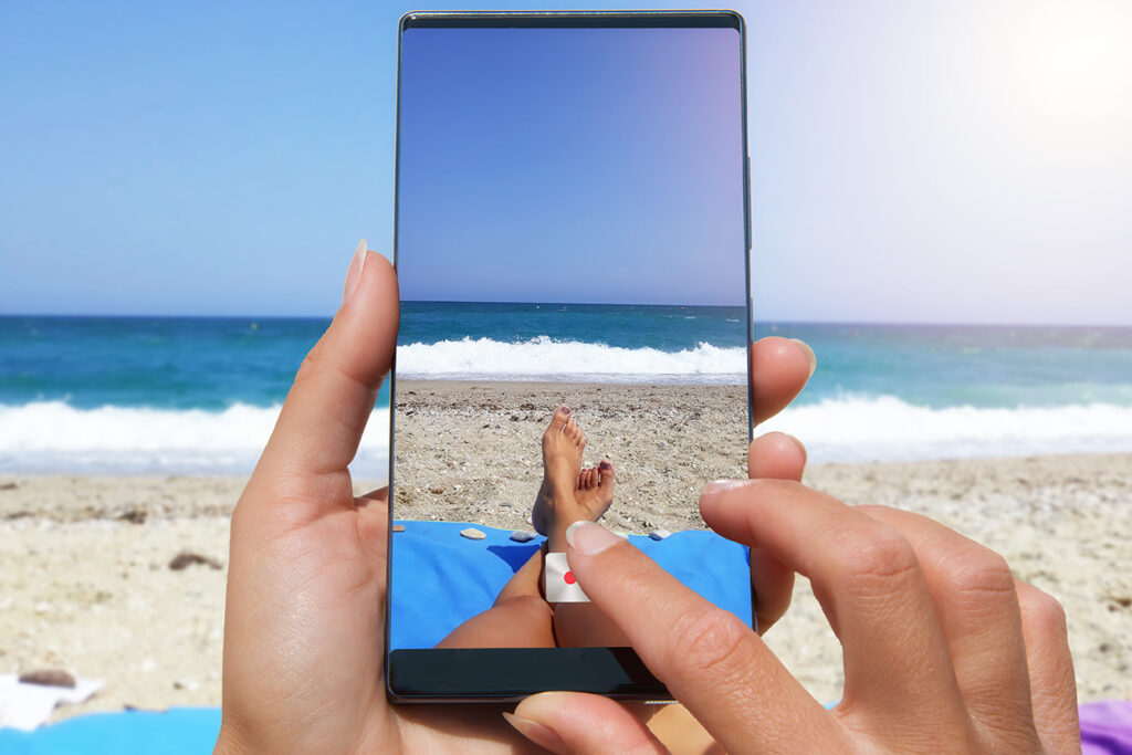 Close-up of hands filming sandy beach and ocean view with a smartphone while relaxing on a beach towel.