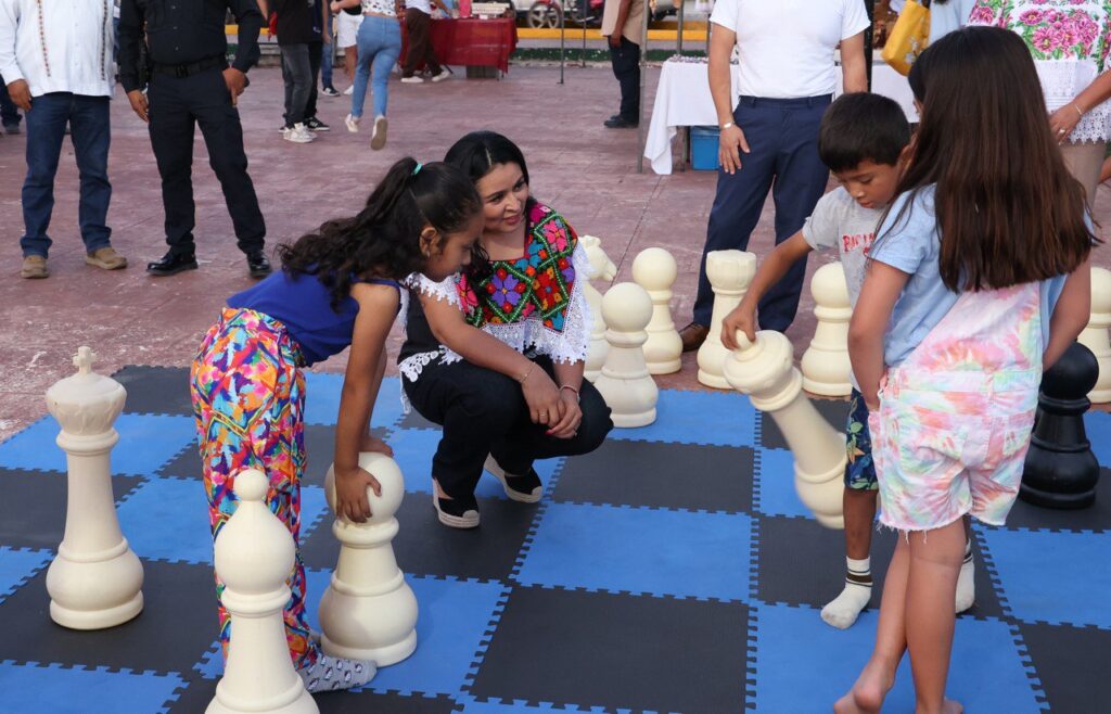 Children play a game of giant chess with oversized pieces as a local official kneels nearby, engaging with them during the Mosaico Cultural event in Leona Vicario.