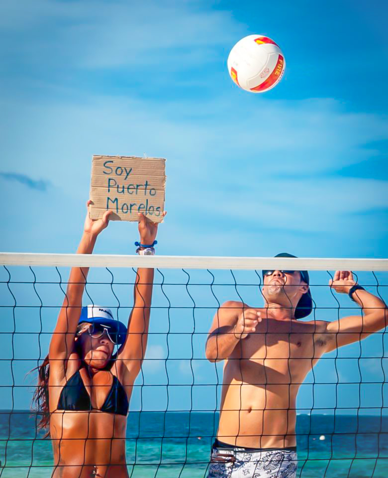 Young adults playing beach volleyball in Puerto Morelos, with a sign reading “Soy Puerto Morelos”