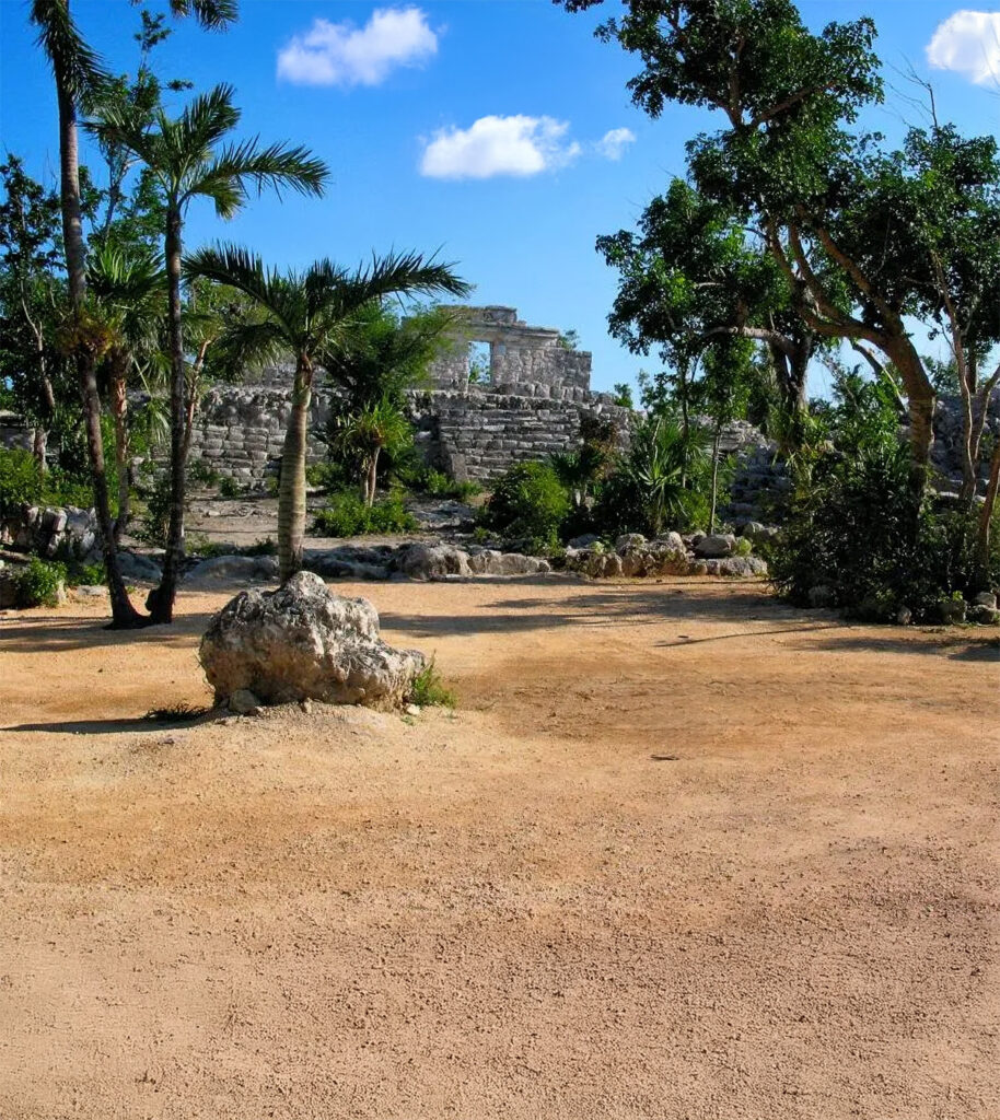 Restored Maya ruins at Xcaret with palm trees and open sandy ground under a bright blue sky.
