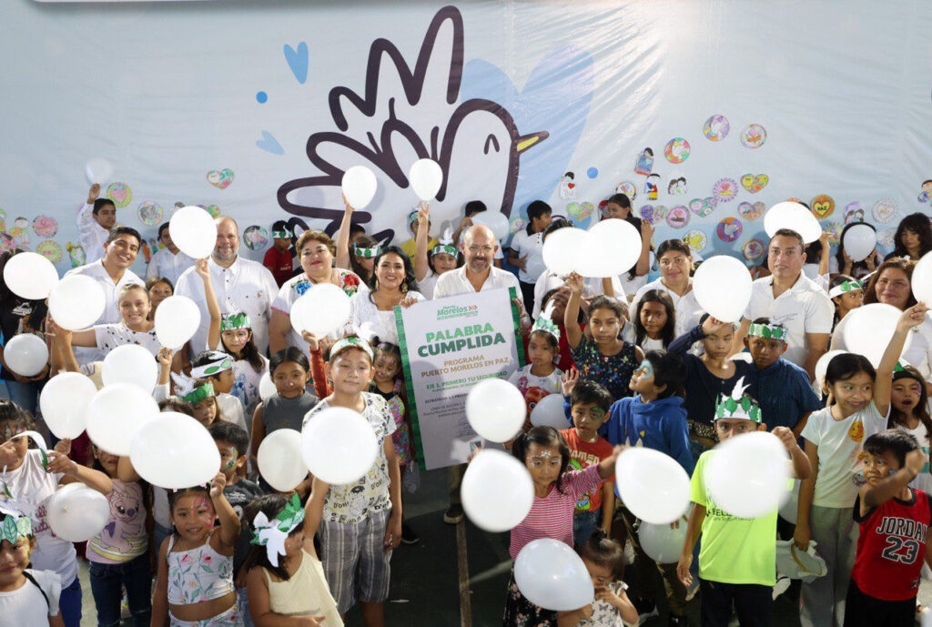 Group of children and officials holding white balloons in front of peace dove mural