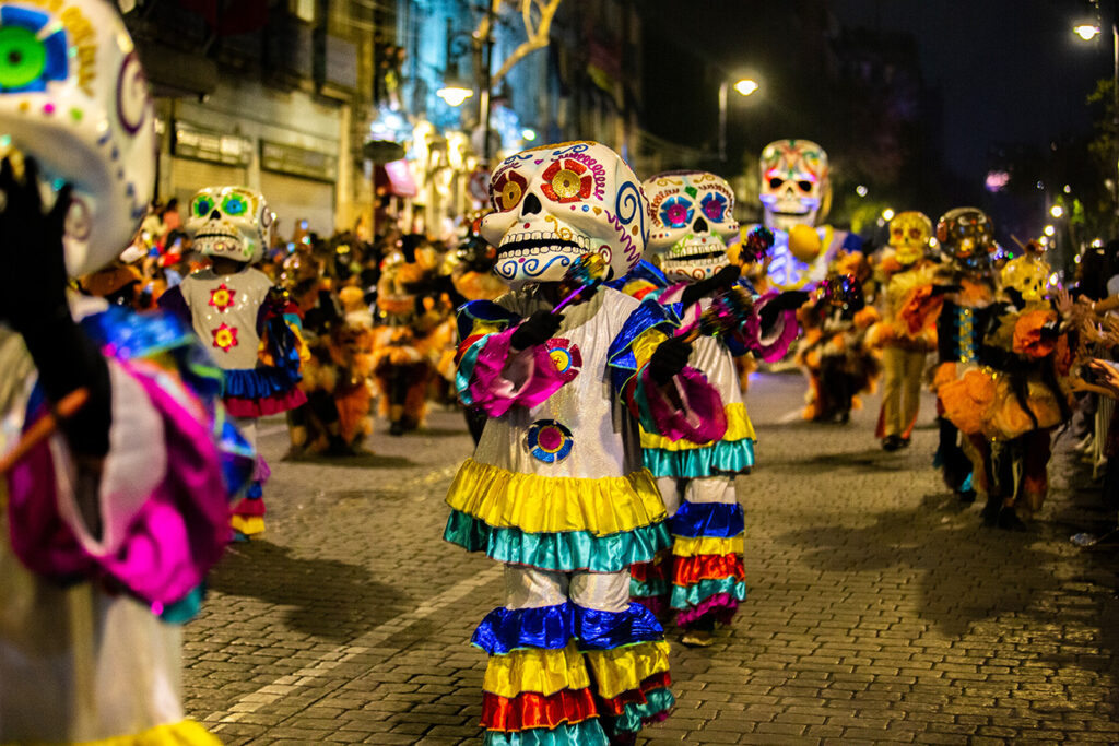 This photograph captures a performer participating in a Janal Pixán celebration in Puerto Morelos, wearing a detailed costume adorned with bright marigold flowers, feathers, and skeletal motifs. The attire represents the fusion of Maya and Mexican traditions during the annual Food for the Souls festivities, symbolizing respect for ancestors and the joyful remembrance of life and death.
