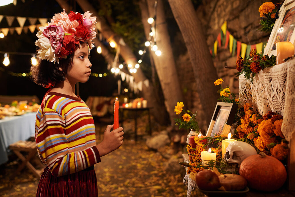 Young girl holding a candle beside a decorated Janal Pixán altar with flowers and candles.