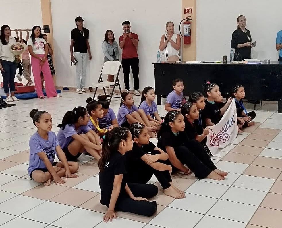 Young gymnasts sit on the floor listening during the Puerto Morelos Artistic Gymnastics Cup while parents watch nearby.