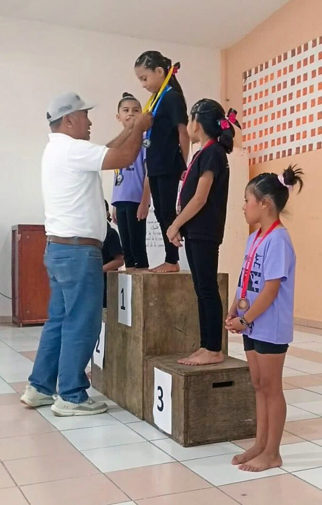 Coach awards medals to young gymnasts standing on the podium during the Puerto Morelos gymnastics competition.