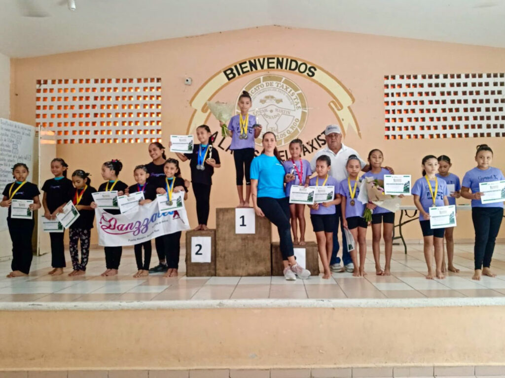 Young gymnasts and coaches pose on stage with medals and certificates after the Puerto Morelos Artistic Gymnastics Cup.
