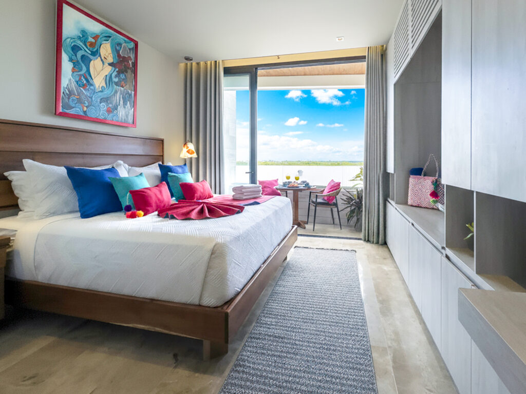 Bright studio with king bed, built-in storage, and a balcony with outdoor seating and blue-sky views.