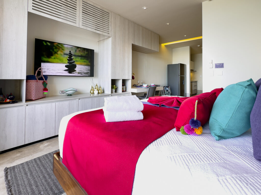 Bright studio interior with king bed, colorful pillows, and built-in cabinets facing a flat-screen TV.