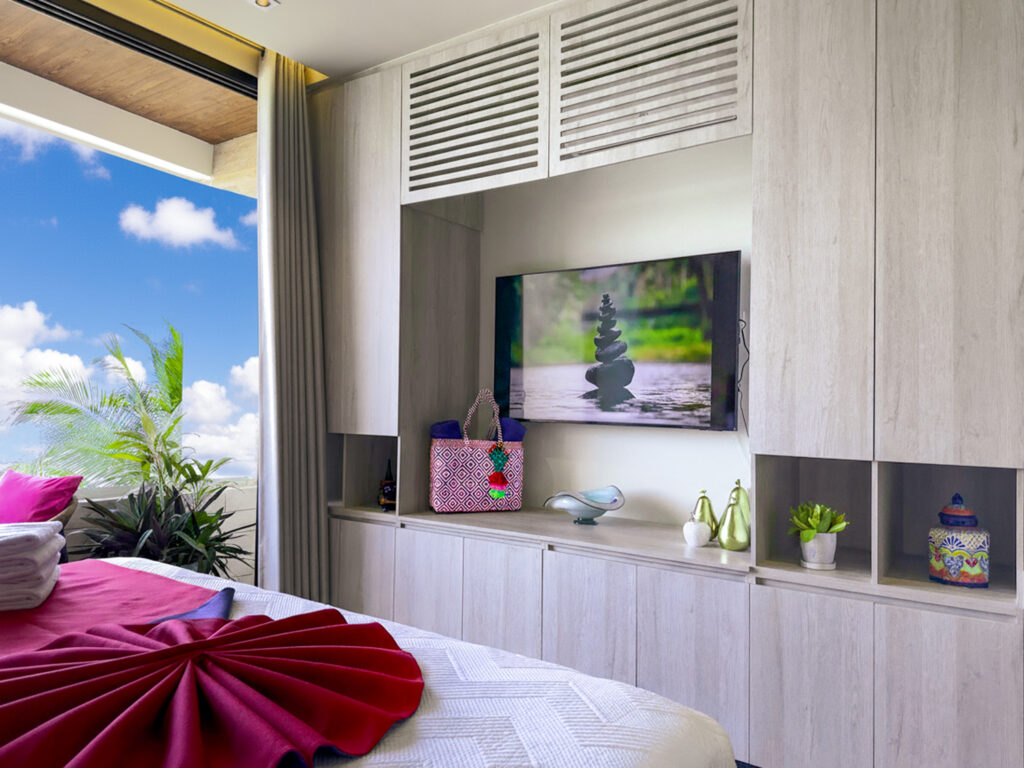Modern studio interior with built-in cabinets, flat-screen TV, and balcony view with blue sky.