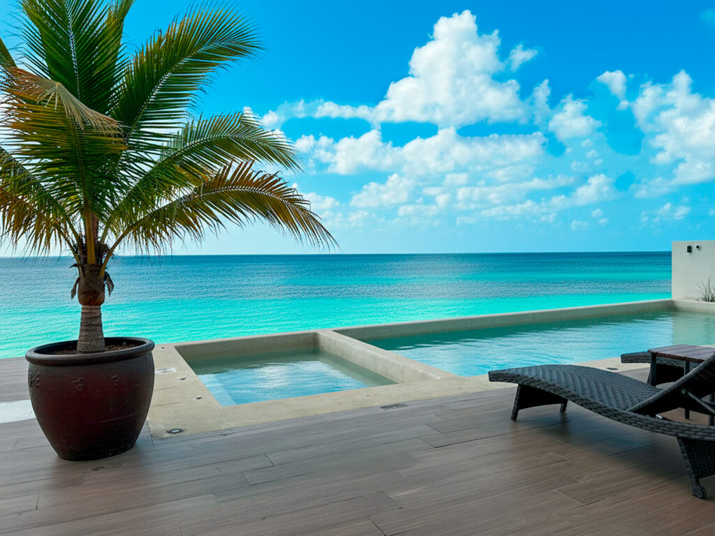 Beachfront pool with turquoise water, palm tree, and ocean view under a bright blue sky.