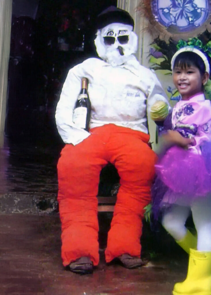 Child standing beside an “Año Viejo” effigy with a painted skull face and bright clothing, ready for a New Year tradition.