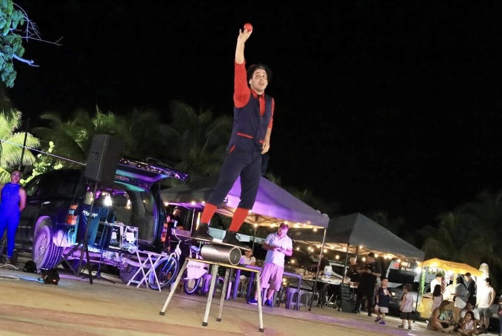 Performer stands balanced on stacked chairs at night, one arm raised, with lights and music gear behind.
