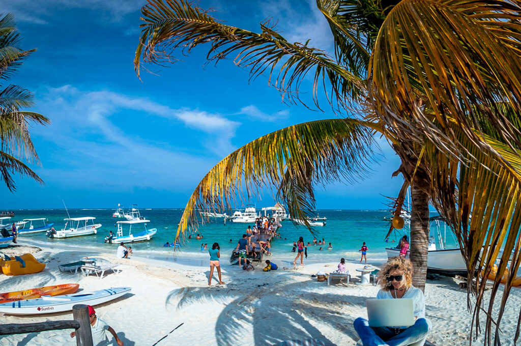 People gather on the beach near the Puerto Morelos pier, with boats, palm trees, and turquoise water.