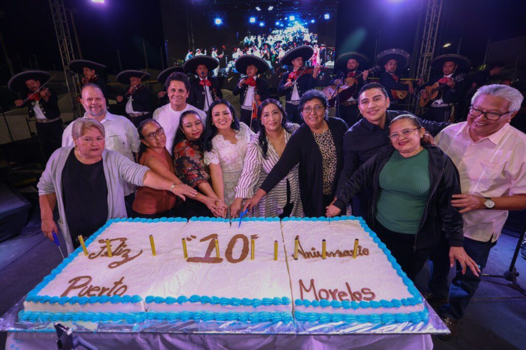 Group smiles behind large 10th anniversary cake for Puerto Morelos at night event.