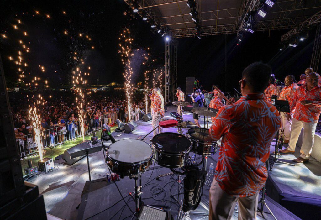 View from behind drummer as band performs on stage with sparks and crowd at night.