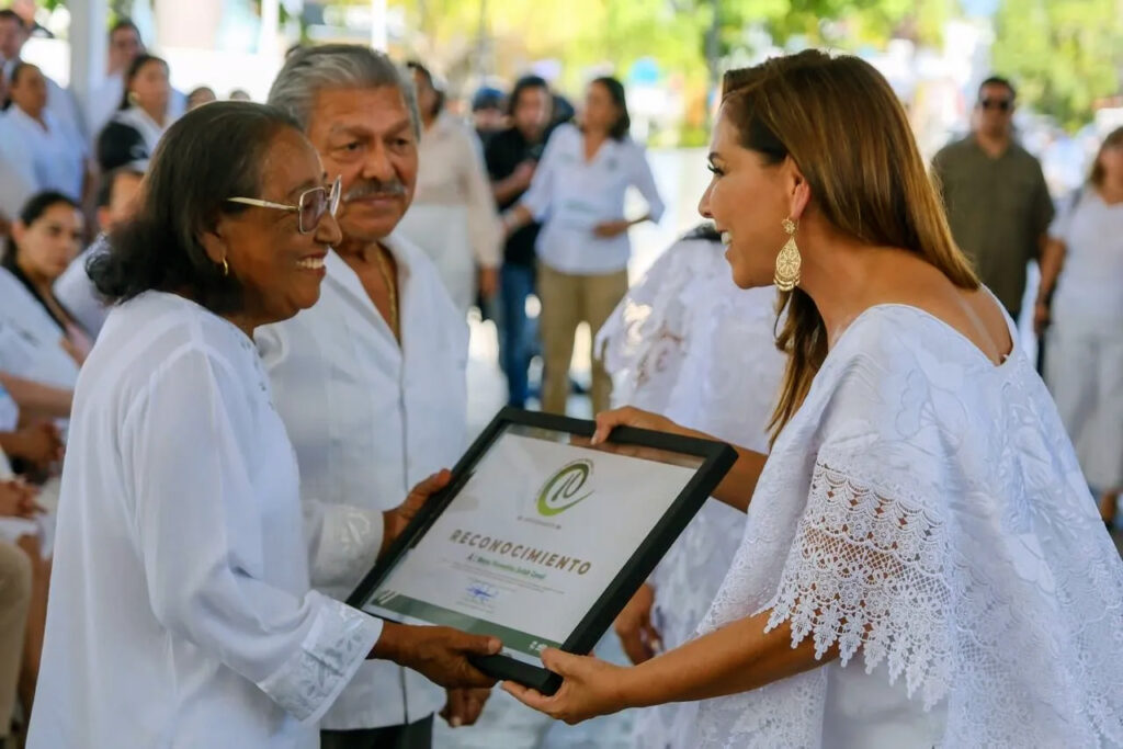 Woman presents framed certificate to another woman at Puerto Morelos anniversary ceremony.