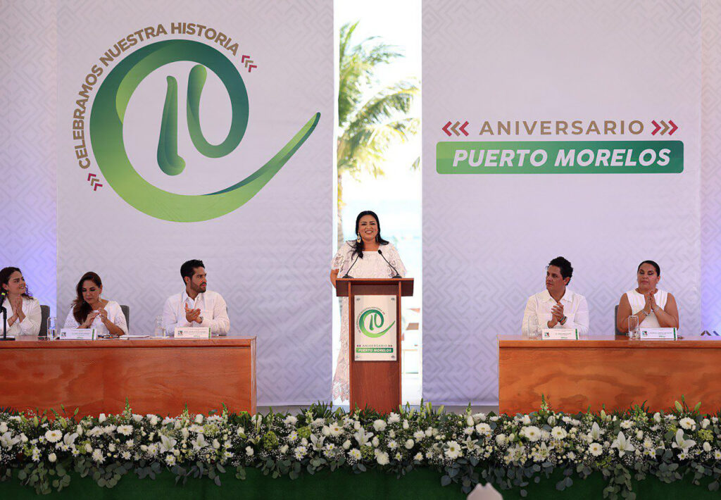 Speaker at podium during Puerto Morelos’ 10th anniversary ceremony, officials seated behind.