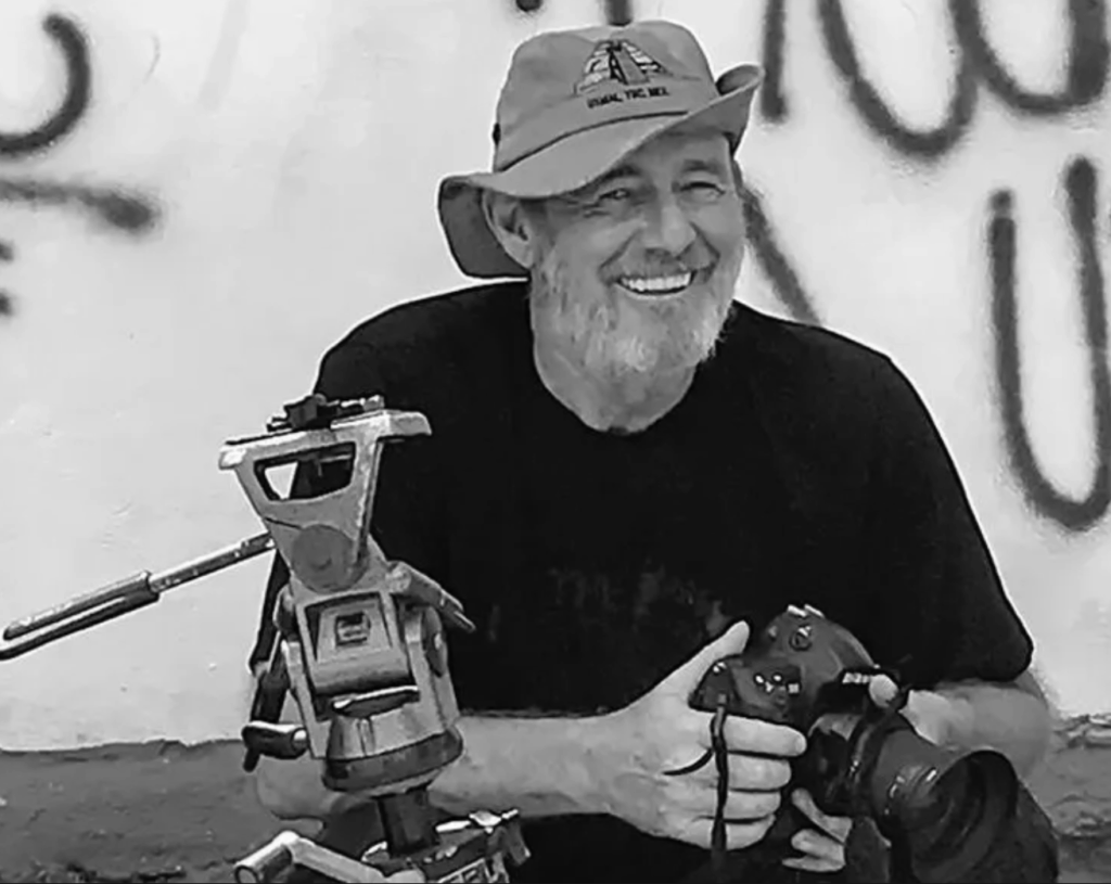 Black-and-white portrait of a smiling photographer holding a camera, with a tripod and background wall behind him.