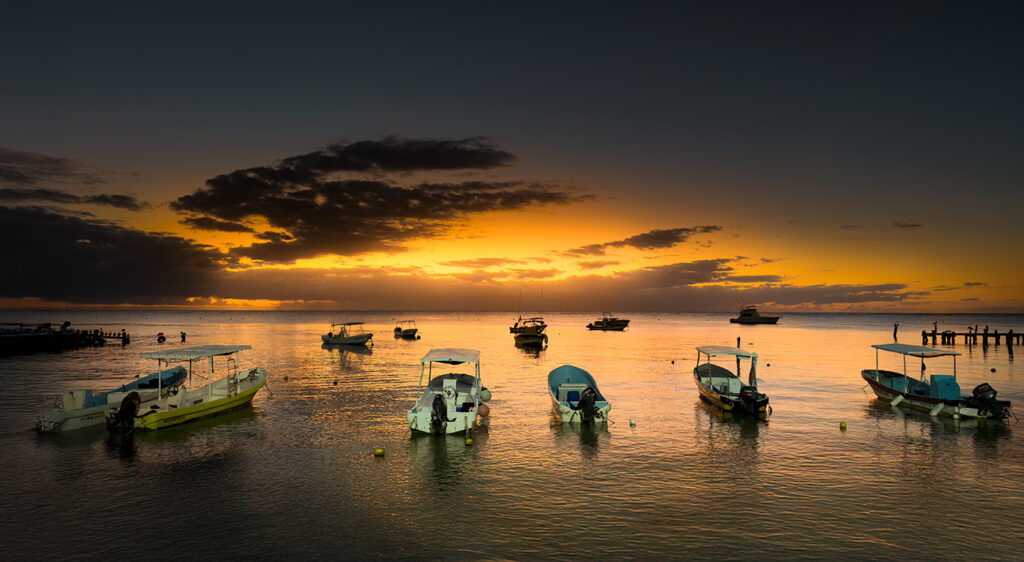 Boats anchored on calm water at sunrise in Puerto Morelos, with a golden sky and low clouds.