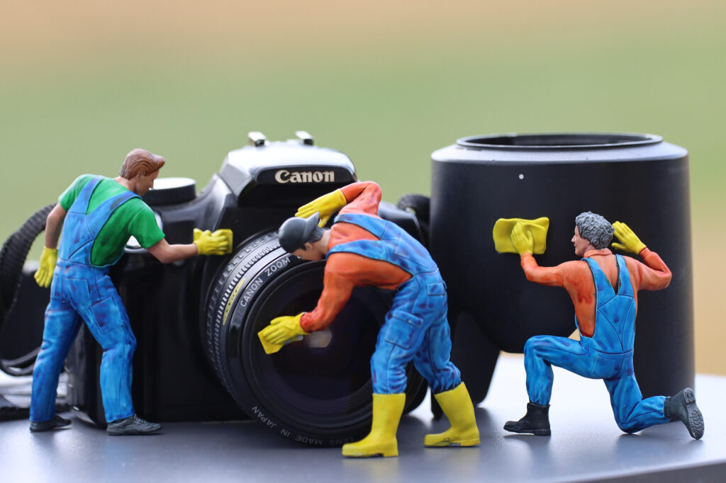 Miniature worker figures “repairing” a Canon camera lens, posed around the lens barrel.