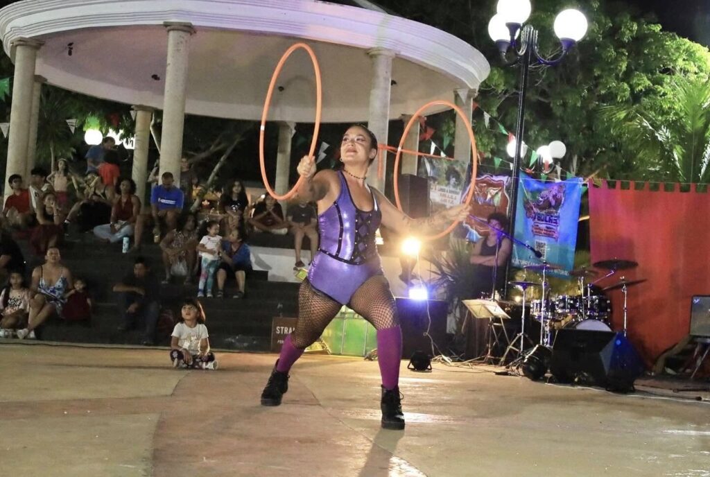 Circus performer spins hula hoops on an outdoor stage as the audience watches in the background.