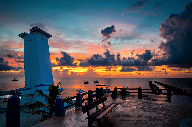 Puerto Morelos lighthouse and pier at sunrise, with colorful clouds over calm water and small boats offshore.