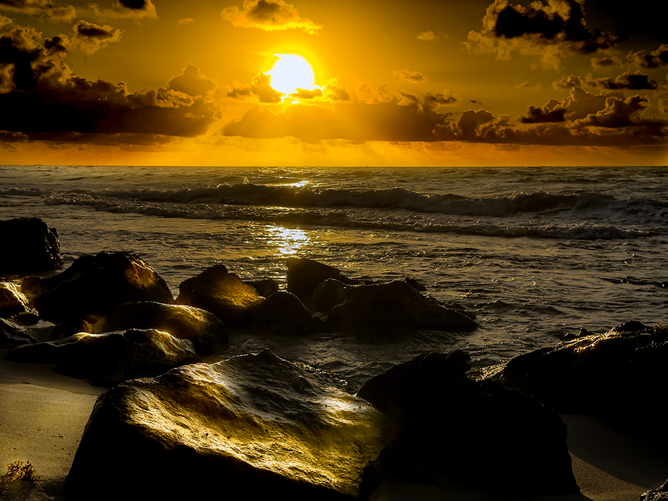 Golden sunrise over the ocean with dramatic clouds, waves, and dark rocks in the foreground.