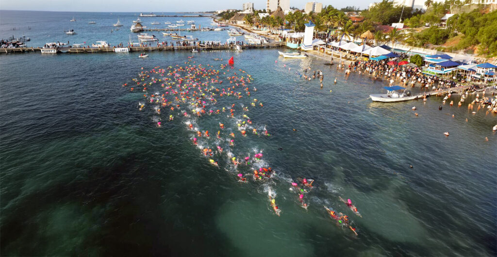 Drone view of dozens of open-water swimmers in colorful caps moving across clear turquoise water near the Puerto Morelos pier and shoreline.